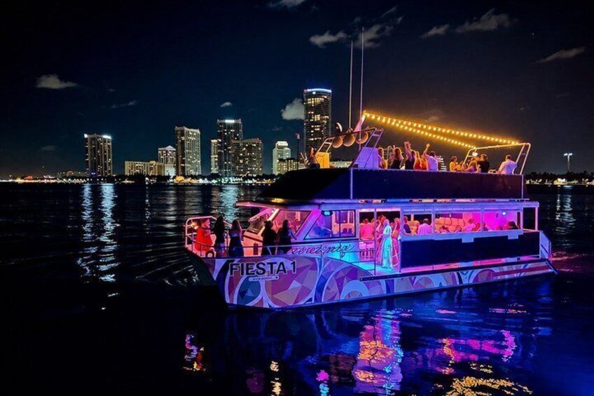 A brightly lit boat with 'Fiesta' written on it, docked at night with a city skyline in the background. 1