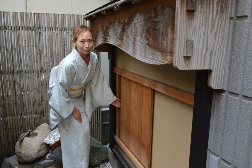 A person in traditional attire standing by a wooden door with a bamboo fence in the background. 2