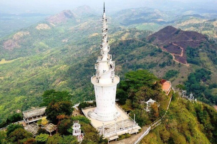 A white tower with a spire, surrounded by lush green hills and a few buildings. 1