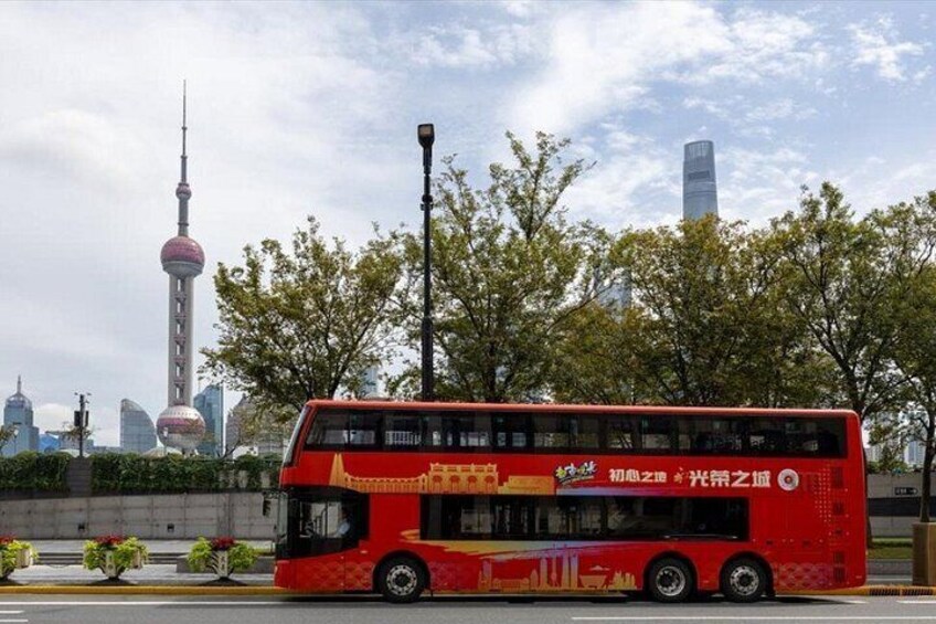 A red double-decker bus with advertisements, parked in front of a modern building with a tower, trees, and a clear sky. 5