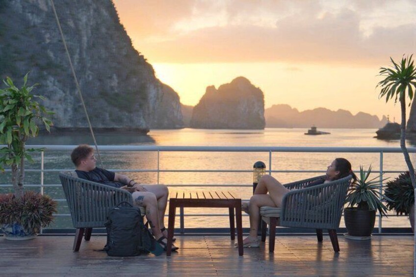 Two people relaxing on wicker chairs by a waterfront with a sunset and rocky cliffs in the background. 1