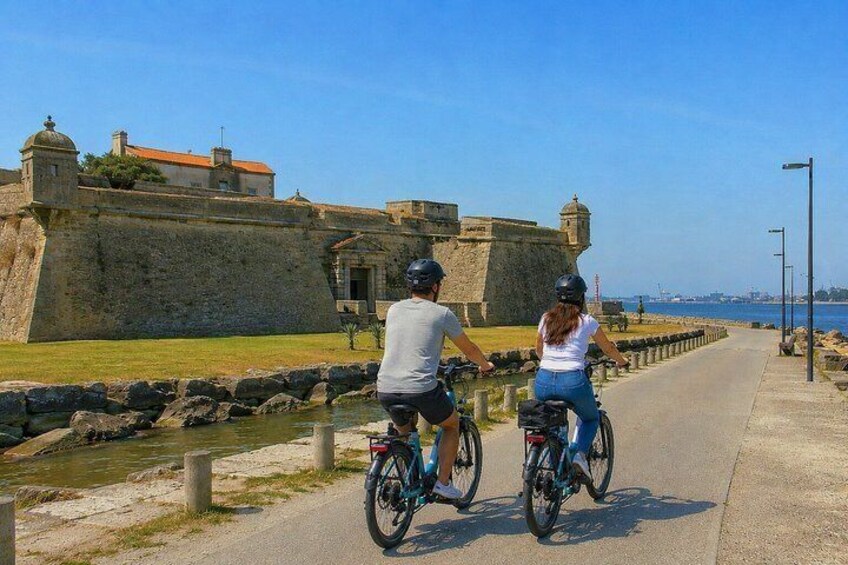 Two people riding bicycles along a paved path beside a body of water, with a historic stone wall and buildings in the background. 5