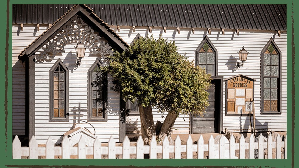 A white church with a black roof, a white picket fence, and a large tree in front. 2