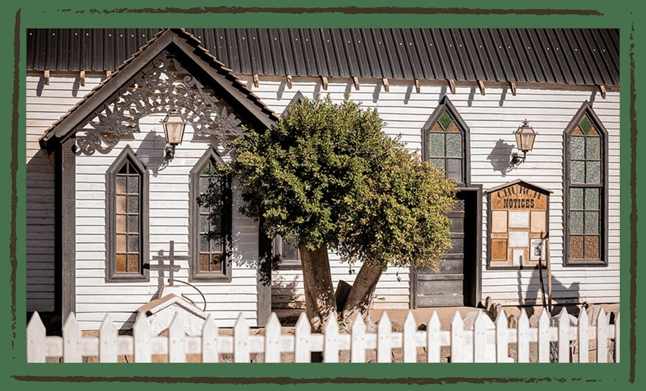 A white wooden building with a black roof, a white picket fence, and a tree in front. 2