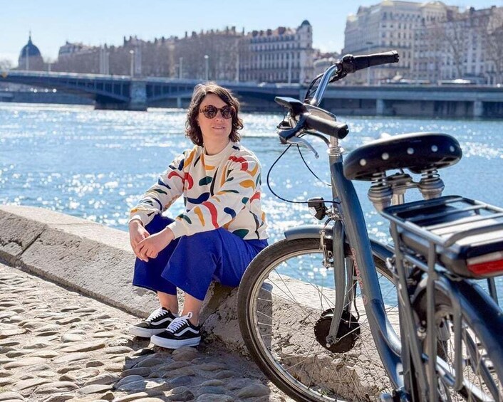 A person sitting on a cobblestone surface next to a bicycle with a cityscape and a river in the background. 1