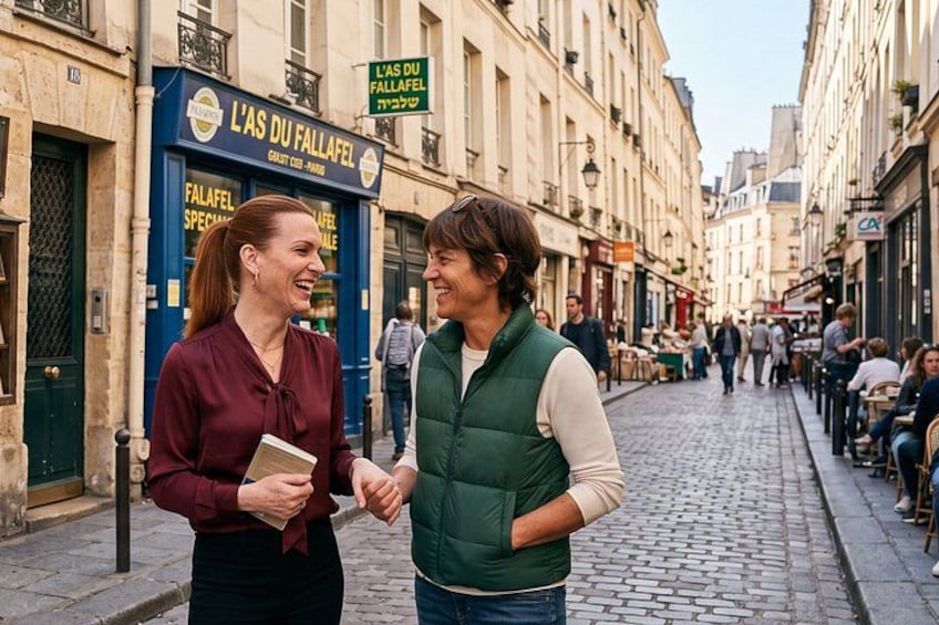 Two people conversing on a cobblestone street with a café named 'L'As Du Fallafel' in the background. 1