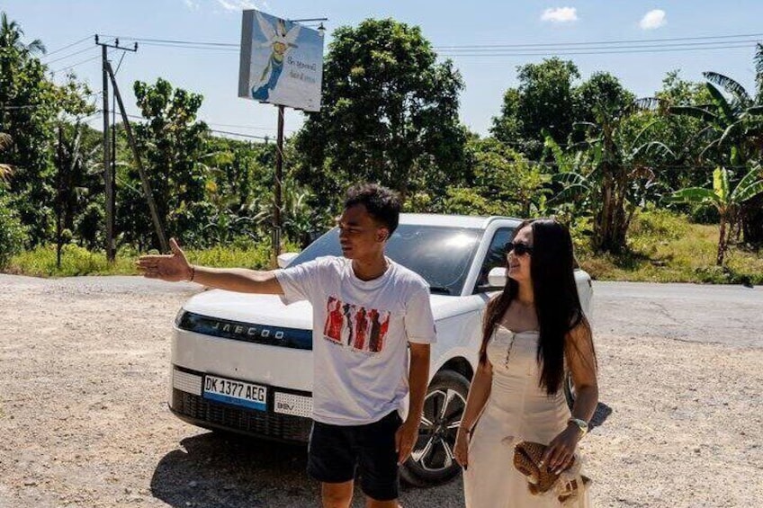 Two people standing next to a white car with a license plate, a billboard in the background, and greenery around. 5