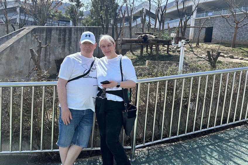 Two people standing near a metal fence with a stone building and trees in the background. 3