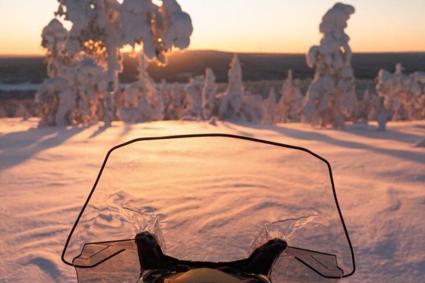 A motorcycle with a clear windshield, set against a snowy landscape with trees and a sunset. 4