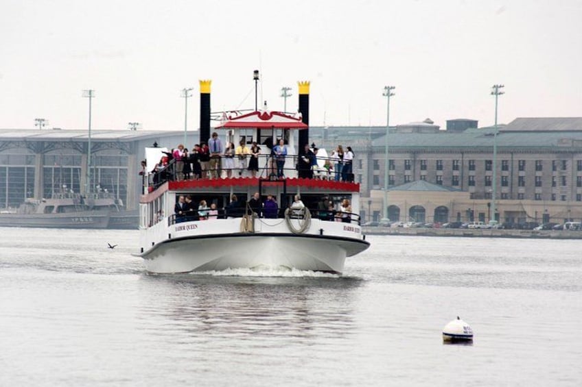 A white boat with red accents and a black funnel, named 'Queen Mary 2,' moving on a body of water with a cityscape in the background. 2