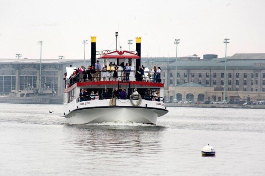 A white boat with a red roof and black funnels, named 'Queen Mary 2,' moving on water with a cityscape in the background. 2