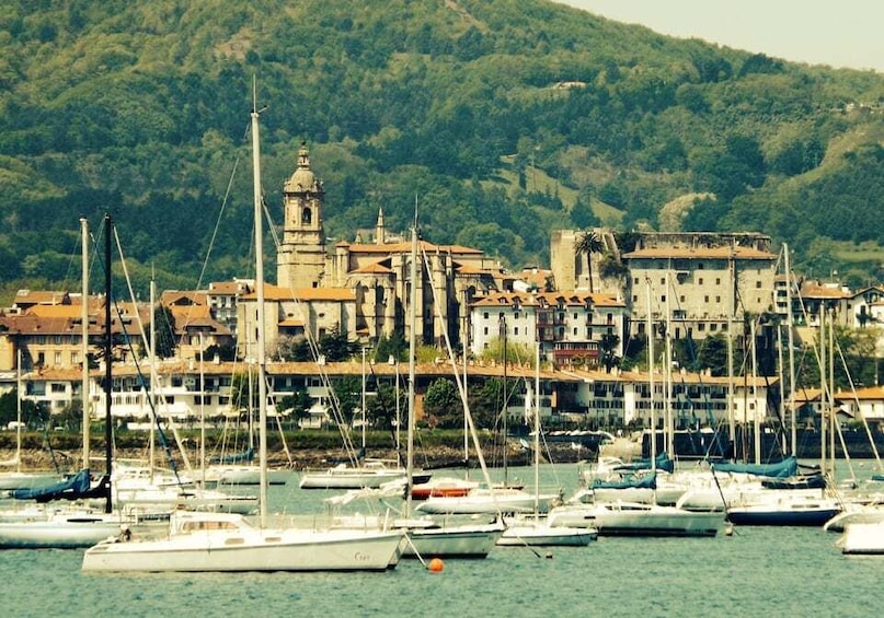 A coastal town with a prominent church tower, a marina filled with sailboats, and a backdrop of lush green hills. 1