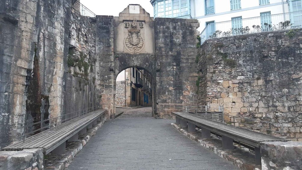 An old stone archway with a coat of arms, leading to a cobblestone street flanked by benches. 5