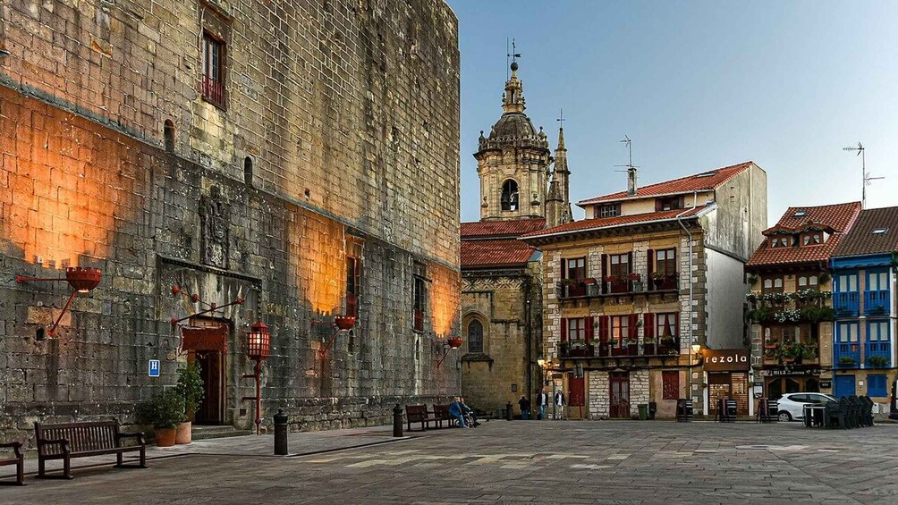 A historic stone building with a bell tower, a street with benches, and a shop named 'rezola.' 3