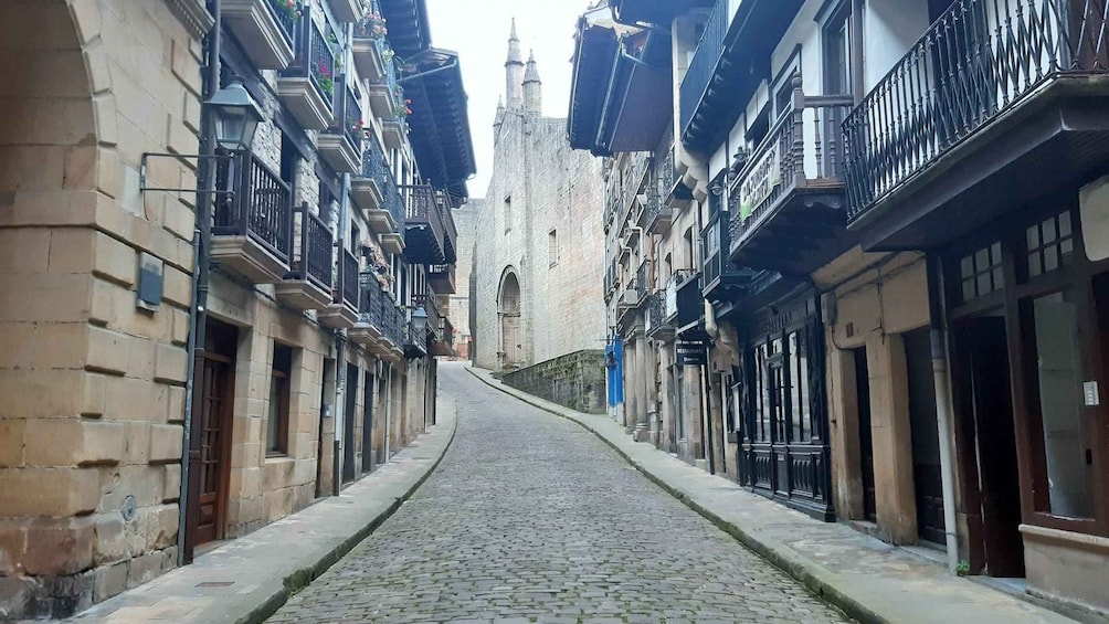 A cobblestone street flanked by historic buildings with balconies and a church steeple in the distance. 2