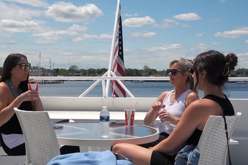 Three people sitting at a table on a boat with an American flag, a body of water, and a dock in the background. 3