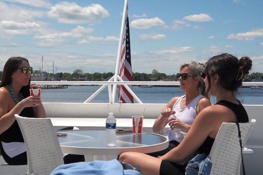 Three people sitting at a table on a boat with an American flag, a body of water, and a dock in the background. 2