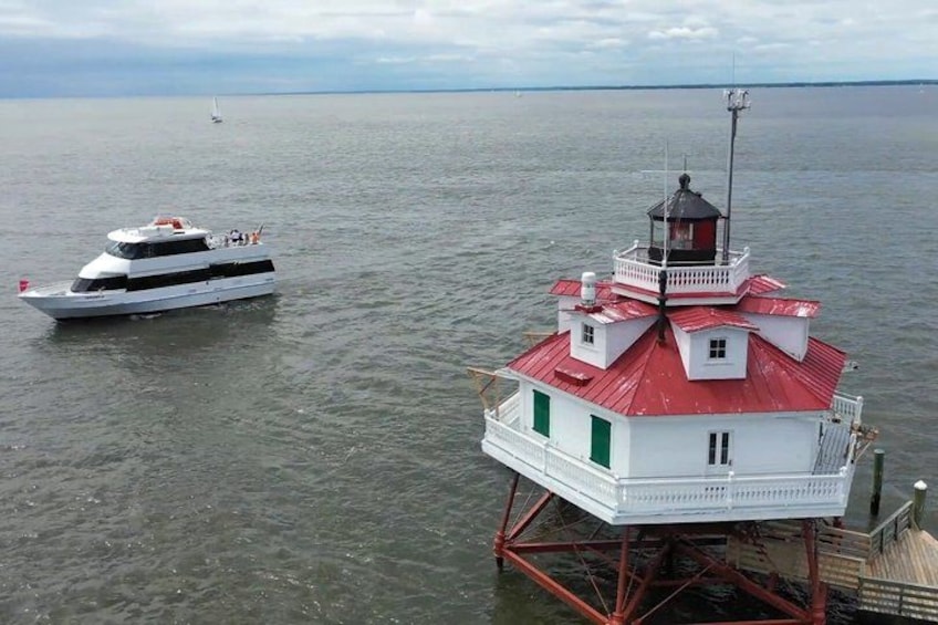 A white boat with a red roof on a calm body of water, a lighthouse with a red roof and white walls, and a distant sailboat. 1