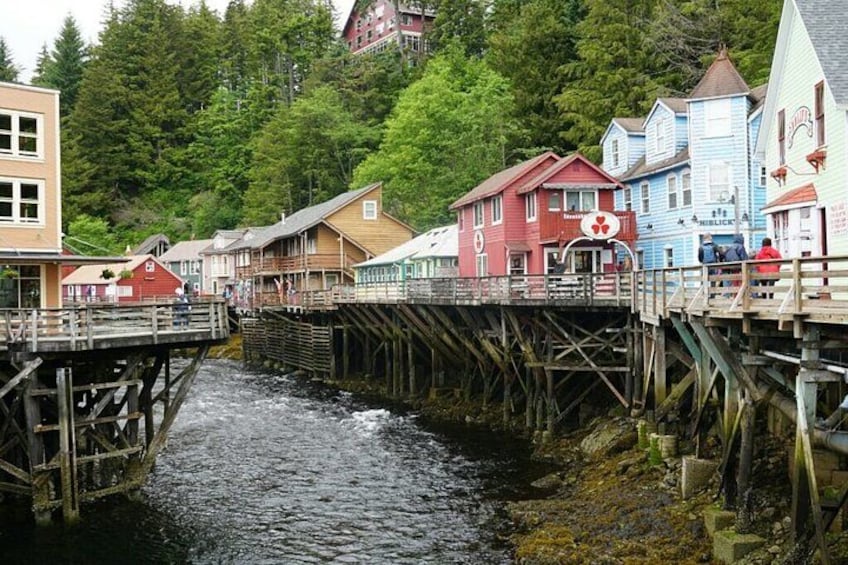 A wooden bridge over a river, colorful houses, and a forested hill in the background. 1