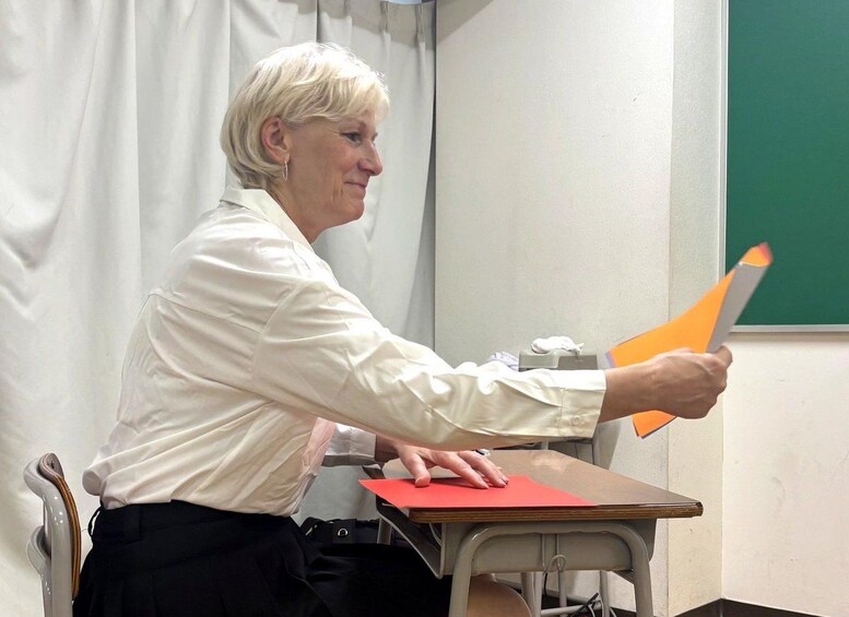 A person holding a large orange folder, seated at a desk with a red folder and a green chalkboard in the background. 3