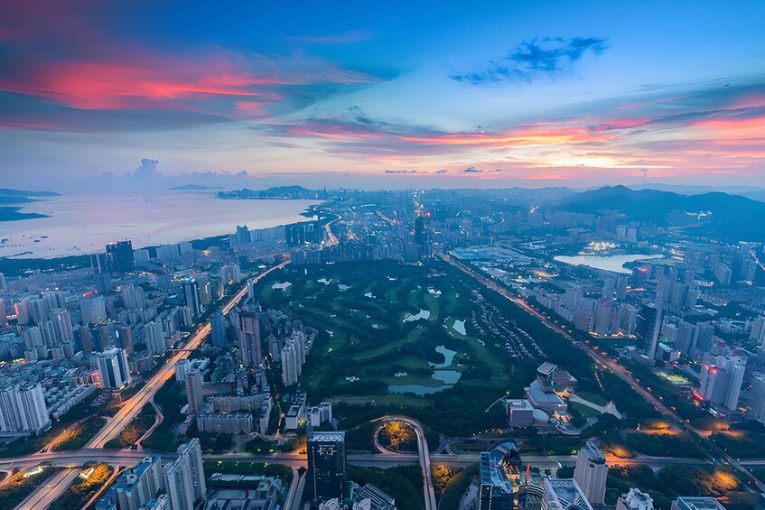 A cityscape at dusk with a river, high-rise buildings, and a park. 1