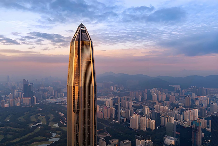 A tall skyscraper with a pointed top, surrounded by a densely packed urban area with various buildings and a distant mountain range. 2