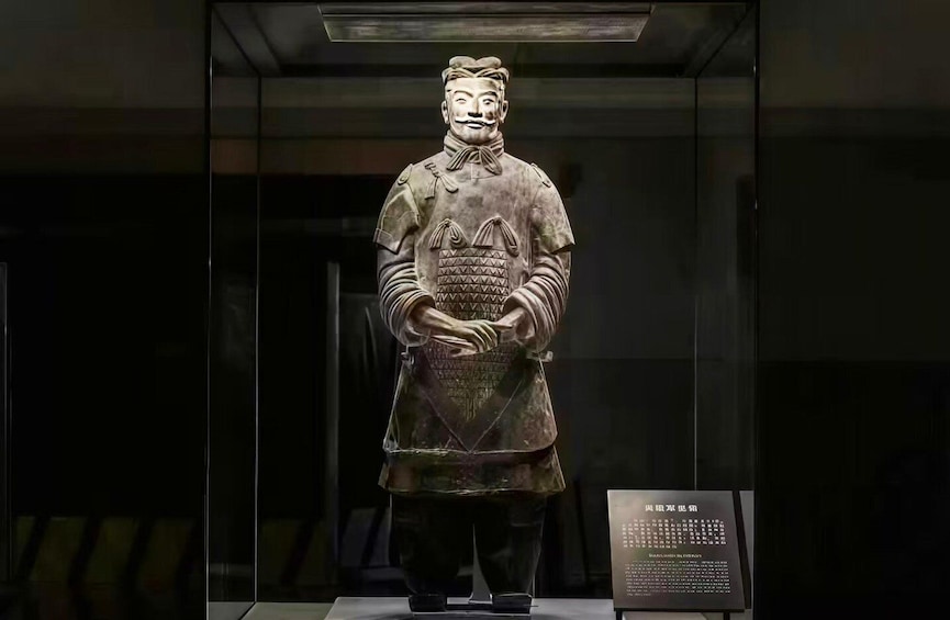 A terracotta statue of a warrior holding a shield and a sword, displayed in a glass case with a placard. 2