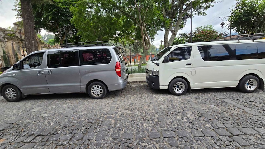 Two parked cars on a cobblestone street with trees and a cross in the background. 1