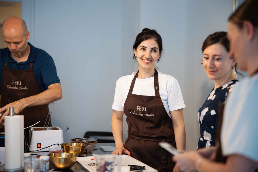 Three people wearing aprons with 'FERI Chocolate Studio' text, working with chocolate bowls and utensils on a table. 3