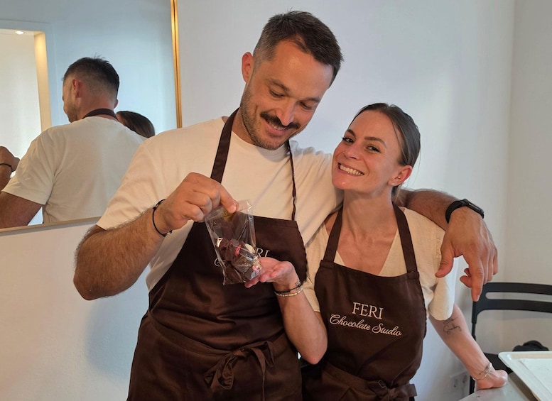 Two people wearing aprons with 'FERI Chocolate Studio' text, holding a chocolate bar and a bottle. 2