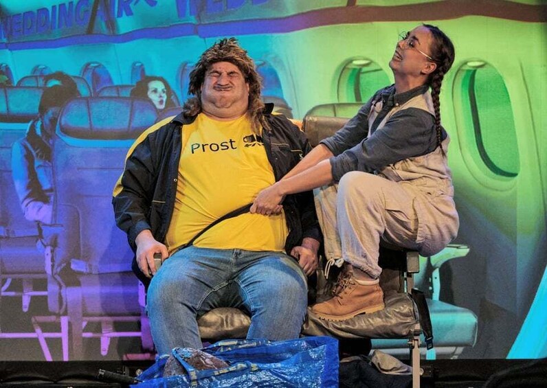 A person in a yellow shirt with 'Prost' on it, sitting on a chair with a bag on the floor. 4