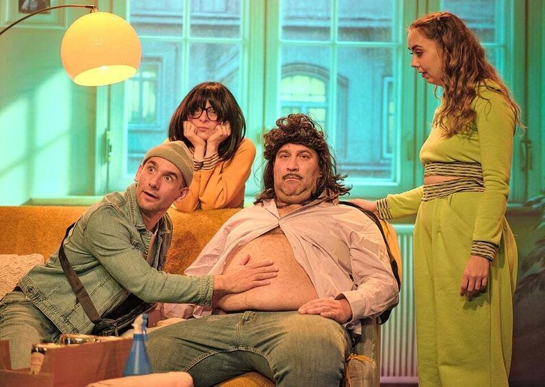 A staged scene with four individuals, one seated and three standing, in a room with a greenish hue and a window in the background. 1