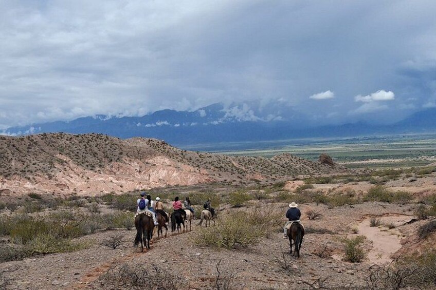 A group of people riding horses on a dirt trail in a desert landscape with mountains in the background. 1