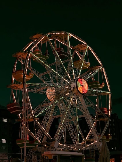 A large Ferris wheel with illuminated seats and a central red light, surrounded by buildings at night. 2