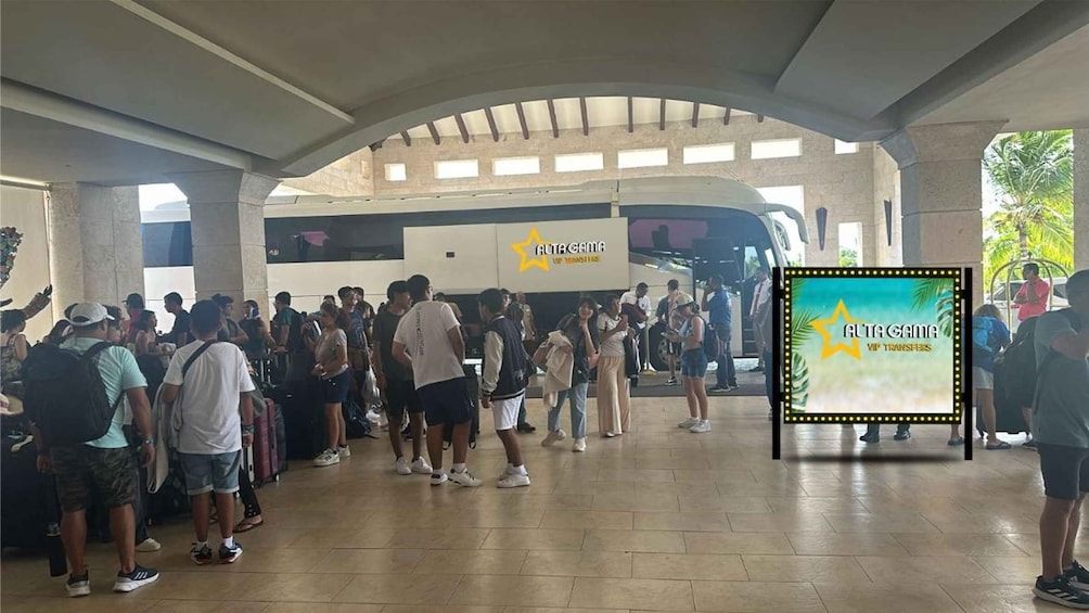 A group of people gathered in a covered area with a bus and a billboard advertising 'ALTA GAMMA VIP TRANSFERS'. 2