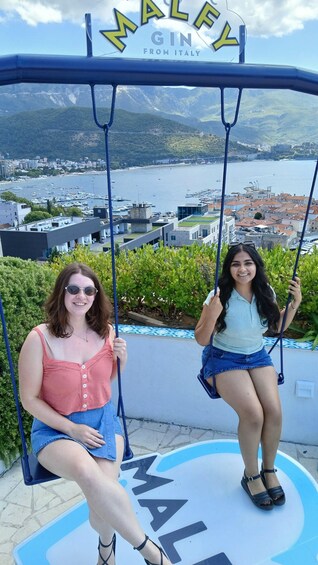 Two women on a swing with a scenic view of a coastal town and mountains in the background. 3