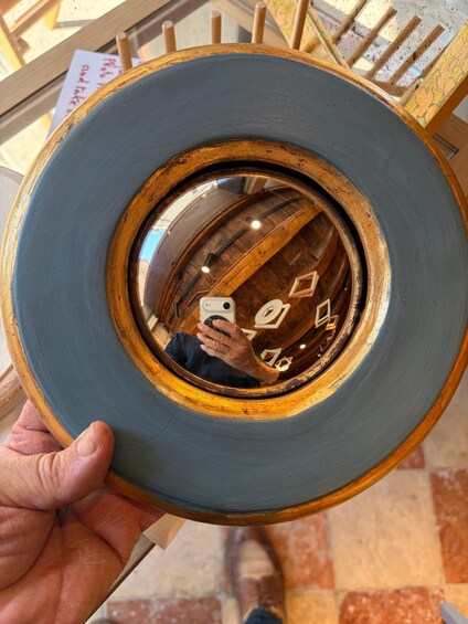 A person holding a circular object with a wooden frame and a dark blue surface, reflecting a person with a smartphone. 3