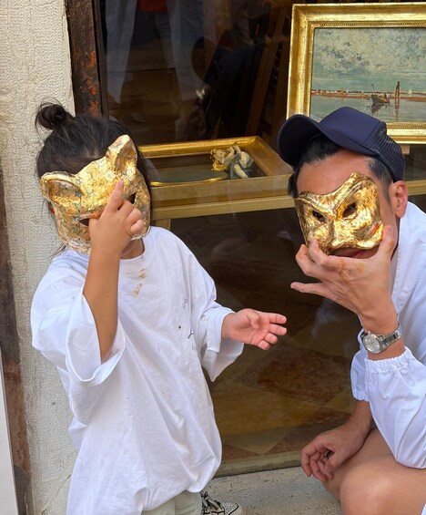 Two people wearing gold foil masks, standing in front of a glass window with a reflection of a painting. 4