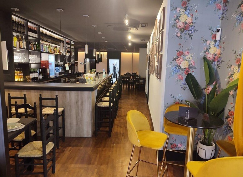 A bar with shelves of bottles, a counter, and chairs. There is a table with a vase and a potted plant. 2