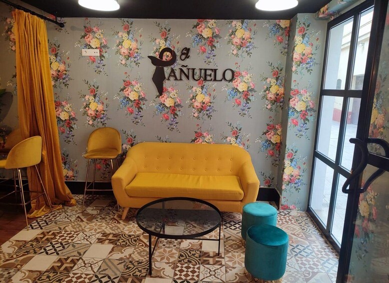 A room with a yellow sofa, two yellow chairs, a round table, and a blue ottoman. The wall has a floral wallpaper and a sign that reads 'El ÁNUELO'. 3