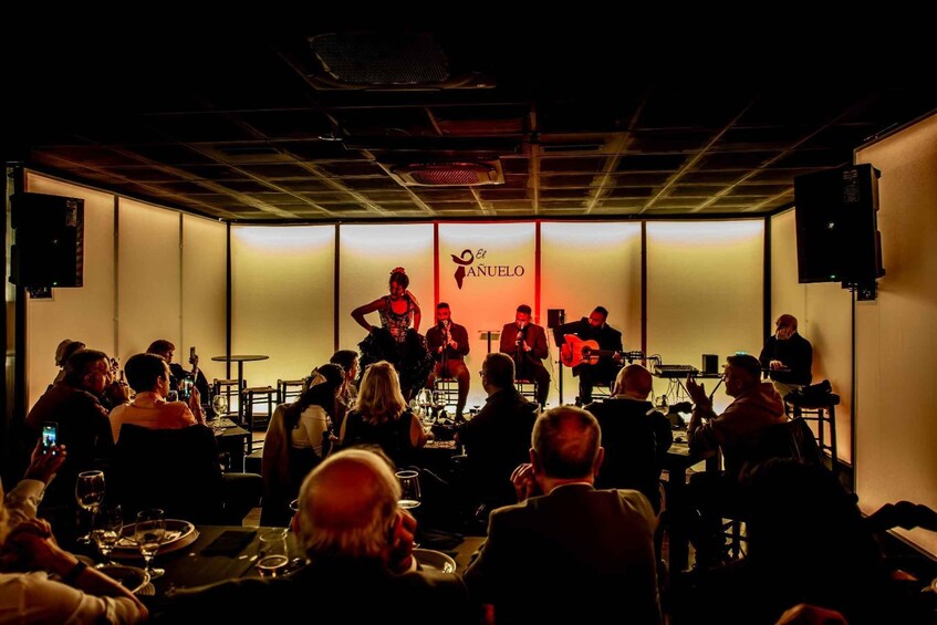 A band performing on stage with a logo that reads 'El Anuelo' in a dimly lit venue with an audience seated in front. 4