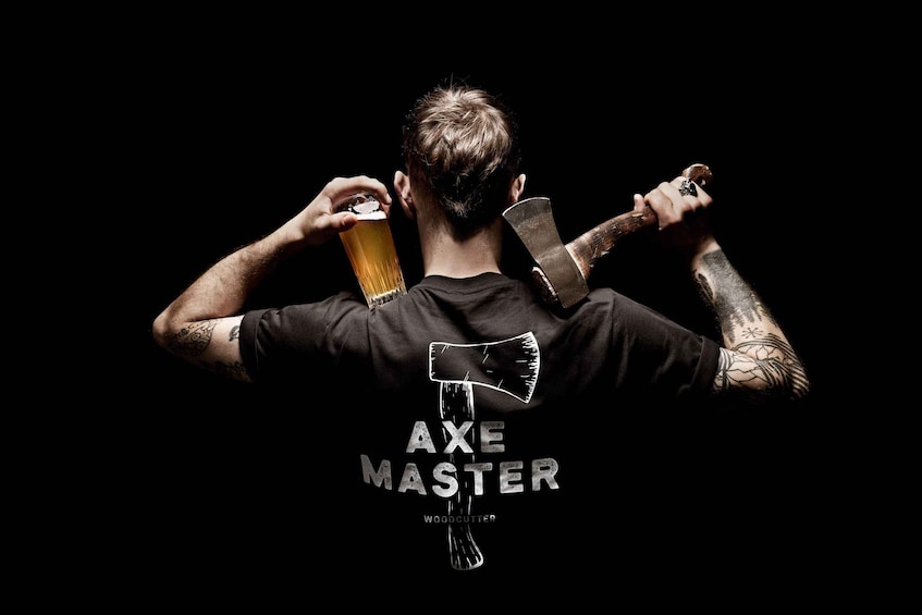 A person with tattoos drinking from a glass, wearing a black t-shirt with 'AXE MASTER' and a wooden axe graphic. 1