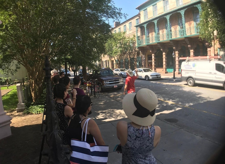 A group of people on a sidewalk, a street with parked cars, and a building with green balconies. 2