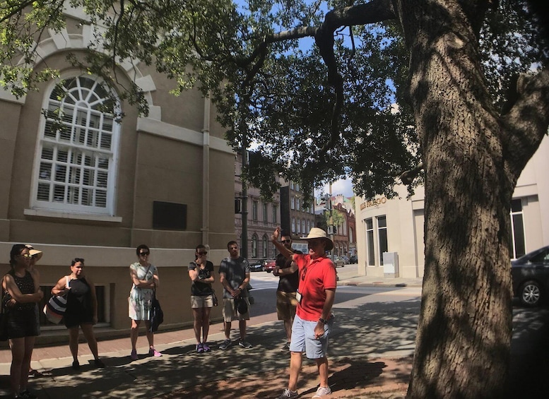 A group of people standing on a sidewalk, a tree, and a building with arched windows. 4