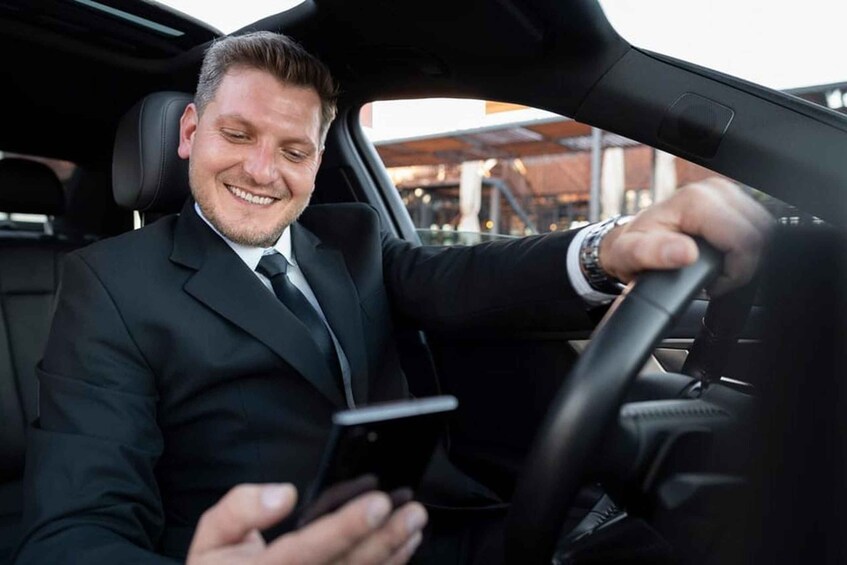 A man in a black suit driving a car, holding a smartphone. 2