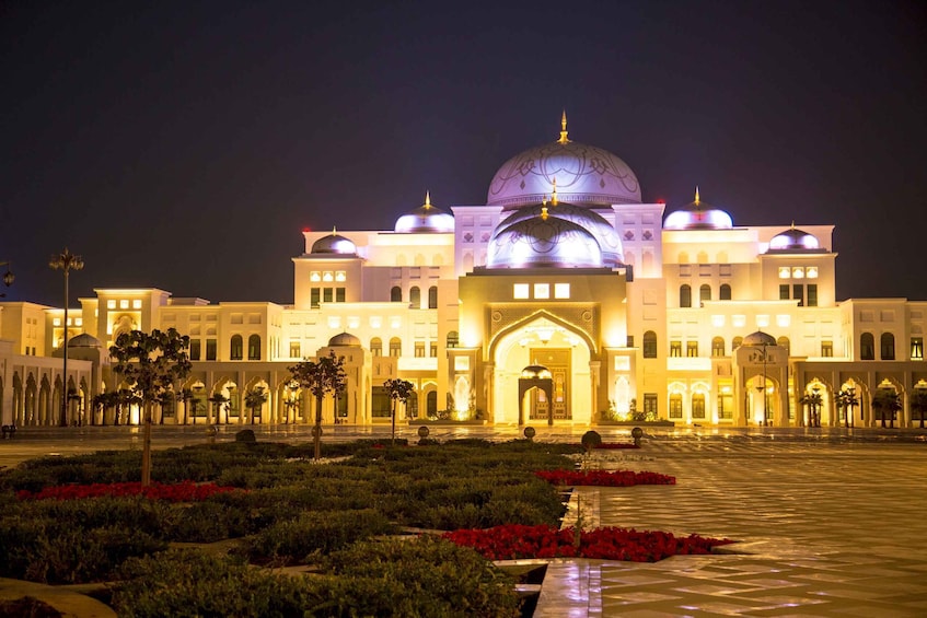 A large, illuminated building with domed roofs and arched windows, surrounded by a well-maintained garden with red flowers. 2