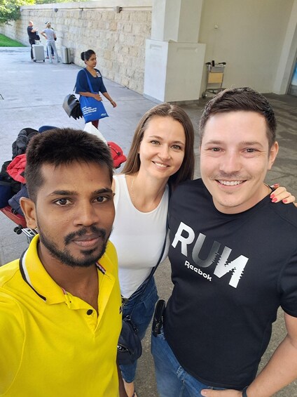 Three people posing for a photo, with a stone wall and a cart with luggage in the background. 1
