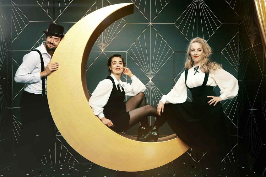Three people posing with a large crescent moon against a geometric patterned background. 1