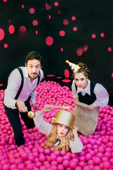 Three people in a ball pit filled with pink balls, with one person holding a golden object. 2