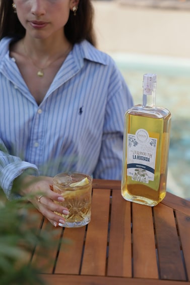 A person holding a glass of vermouth la rugiada next to a bottle of the same drink on a wooden table. 3